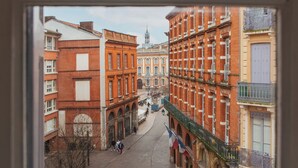 Vista attraverso una finestra su una strada con edifici, tra cui un palazzo in mattoni rossi con un balcone e un edificio giallo con balcone.