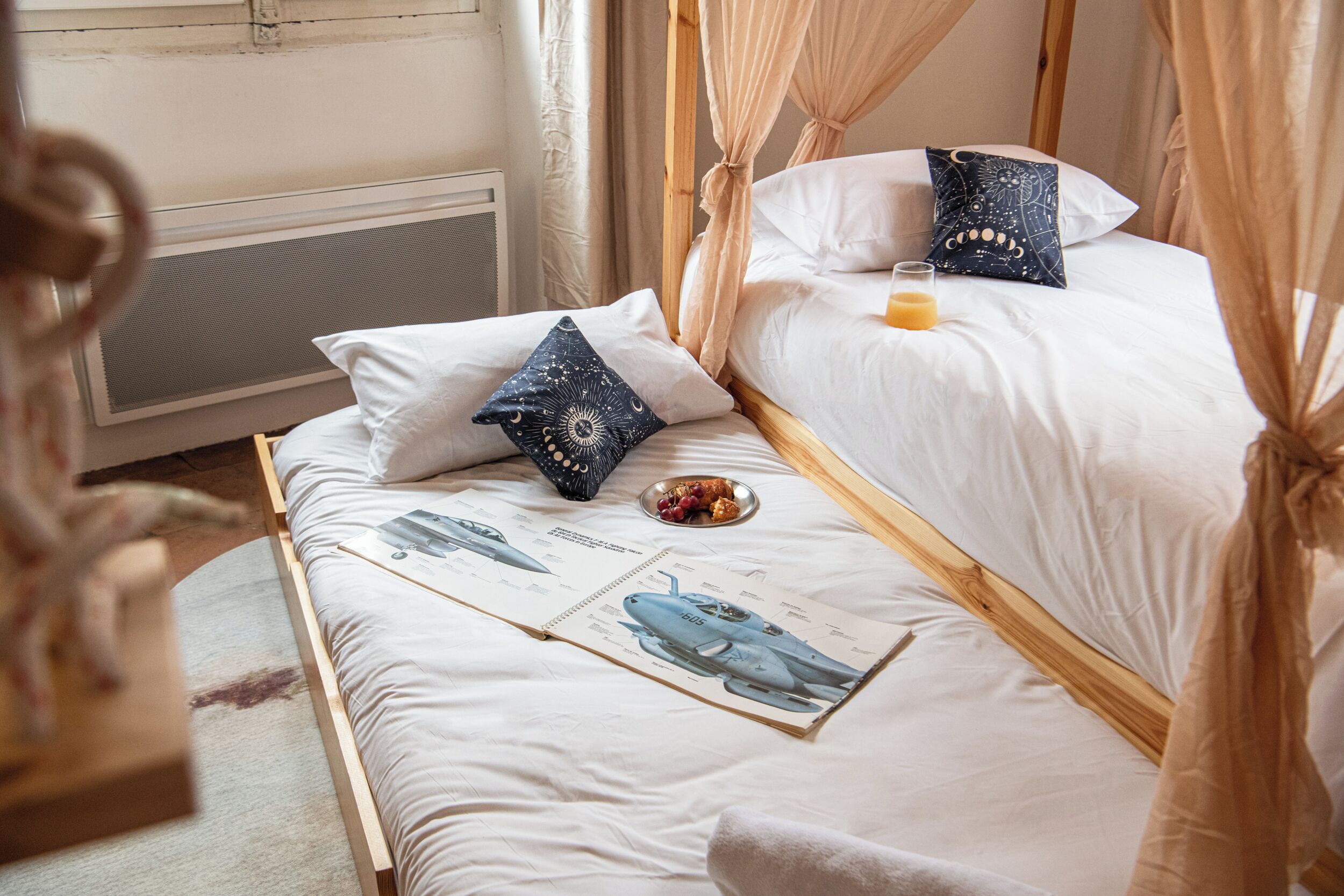 A double bed with a canopy, white bedding, a small bedside table with a glass of orange juice, and a pillow with a starry night design.