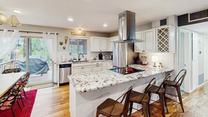 Une cuisine moderne avec des meubles blancs, un îlot central doté de tabourets de bar, une hotte de cuisine en acier inoxydable et une grande fenêtre avec des rideaux.