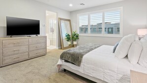 Ein Schlafzimmer mit einem Bett, einem Fernseher auf einer Kommode, einem großen Spiegel und einem Fenster mit Jalousien.
