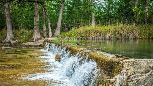 A waterfall cascading over a rocky ledge into a calm river, surrounded by dense forest. - RRR Retreat | 2 Cabins | Medina Riverfront (Medina)