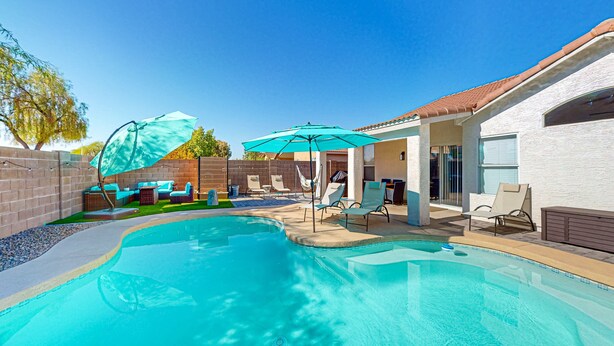 A swimming pool with lounge chairs and umbrellas, surrounded by a brick wall and a white building with a tiled roof. - This charming, single-level home in Chandler is the ideal spot for families (Chandler)