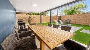 Eine überdachte Terrasse mit einem Holztisch und Korbstühlen, einem Grill und einem kleinen Gartenbereich mit Kunstgrashalm und Ziegelmauern.