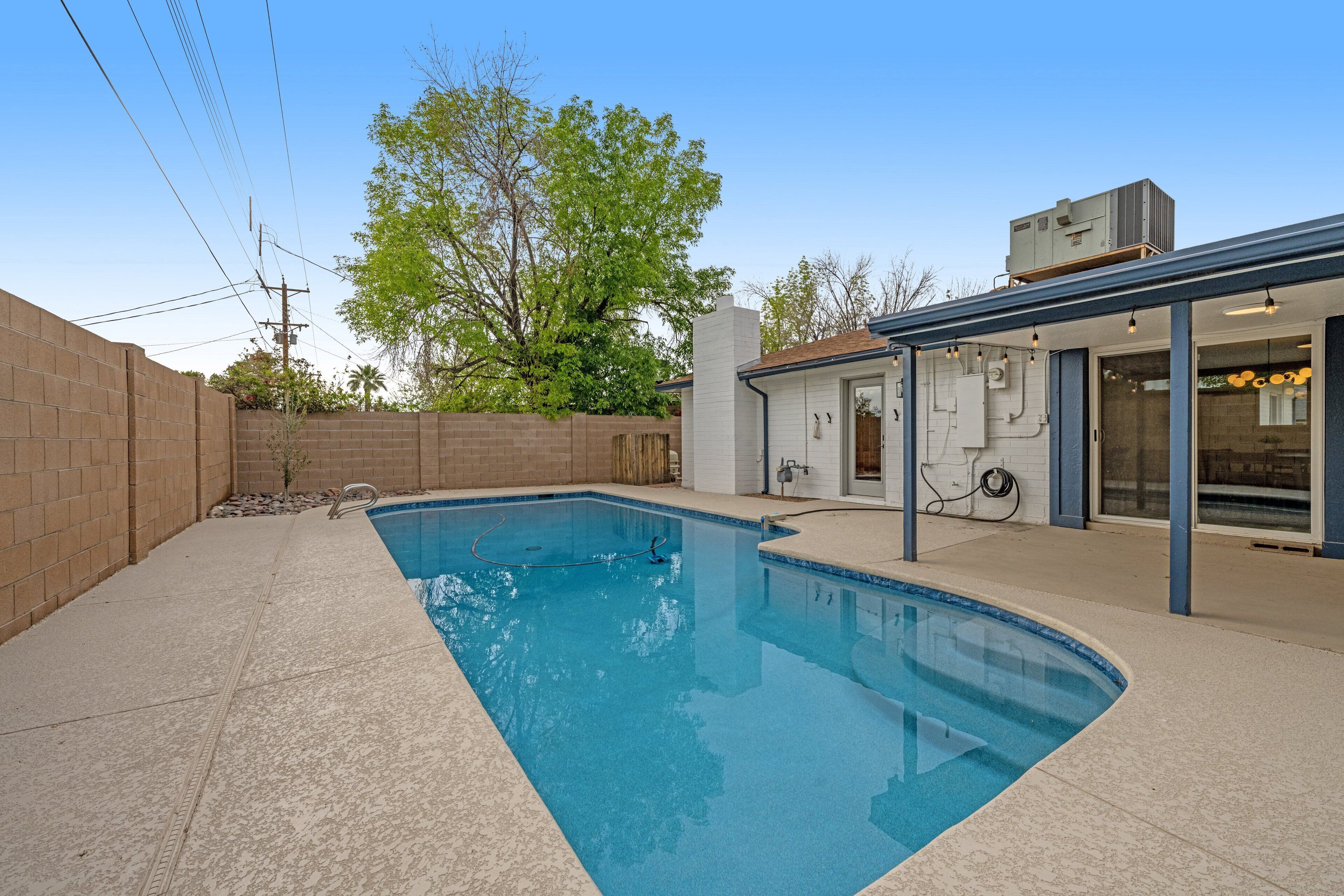 A rectangular swimming pool with a tiled deck, a brick wall, and a covered patio area with a glass door.