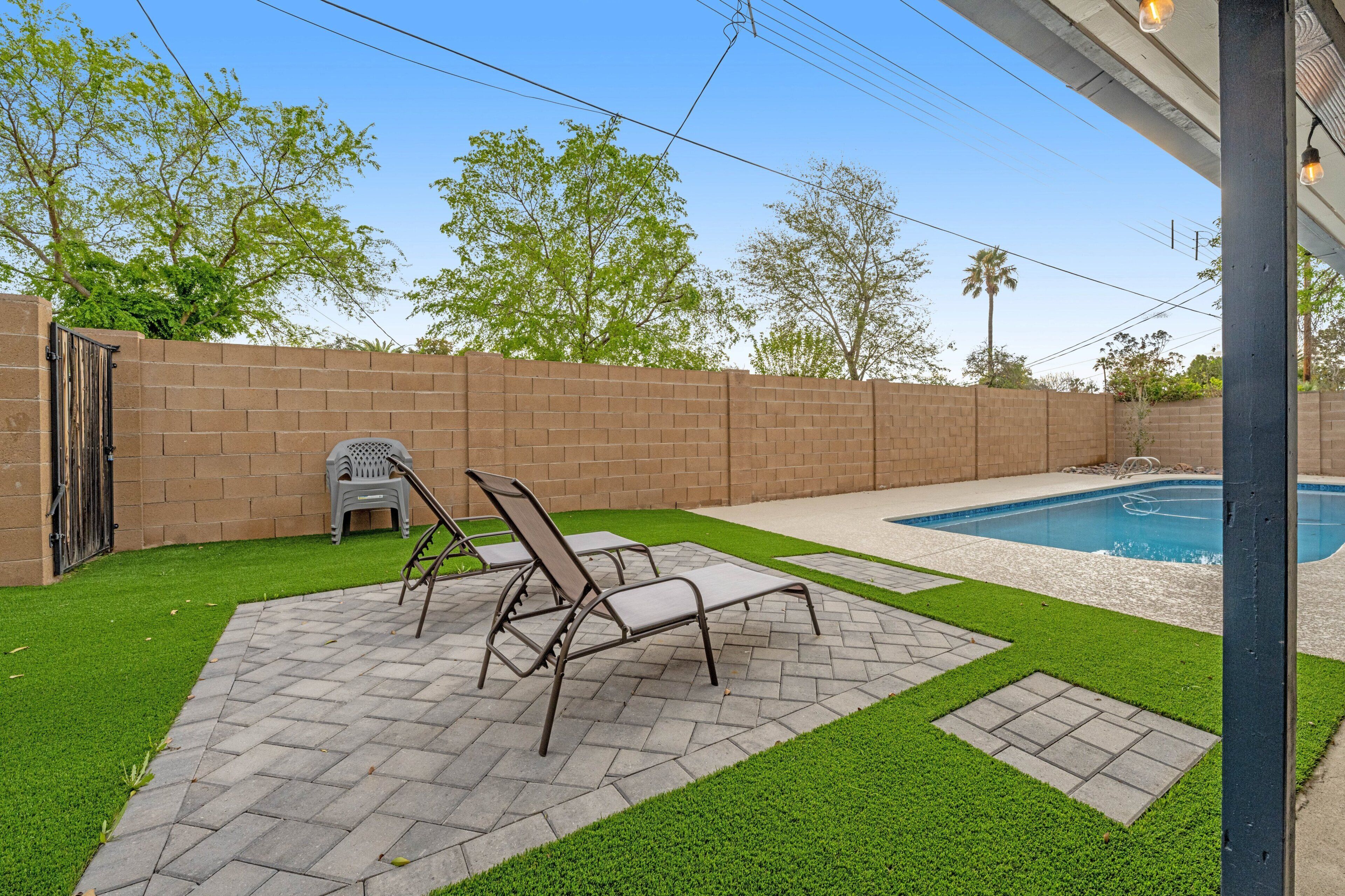 A backyard with a swimming pool, a brick wall, and a patio area with chairs and a small table.
