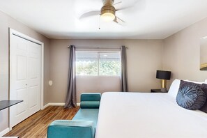 A bedroom with a bed, a ceiling fan, a window with curtains, a bedside table with a lamp, and a small bench. - Dog-friendly condo near attractions with fenced yard, central AC, and cute decor (Glendale)