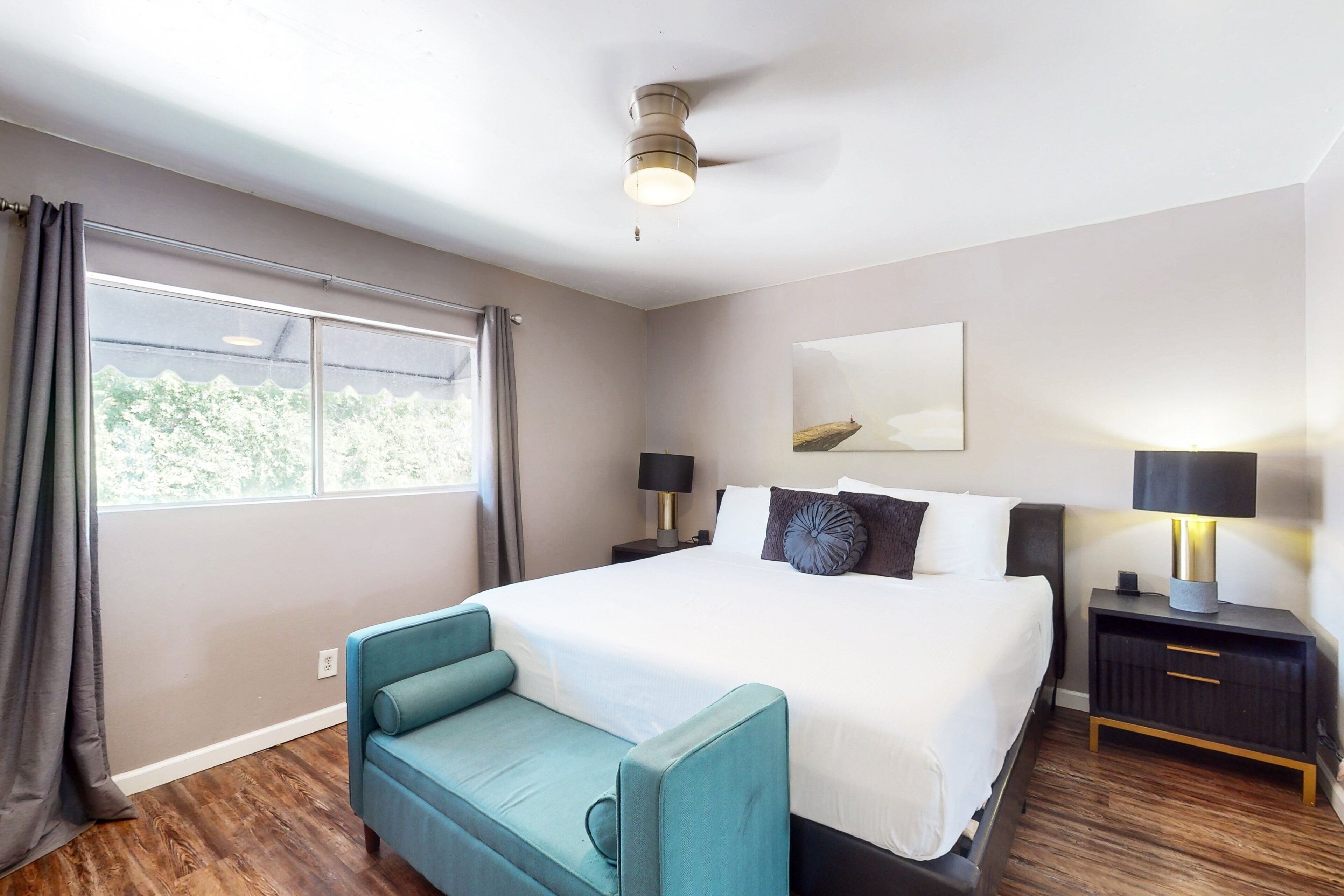 A hotel room with a large bed, two bedside tables with lamps, a window with curtains, a small sofa, and a painting on the wall.