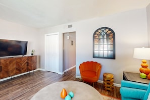A living room with a TV, wooden cabinet, orange chair, blue chair, and a lamp.