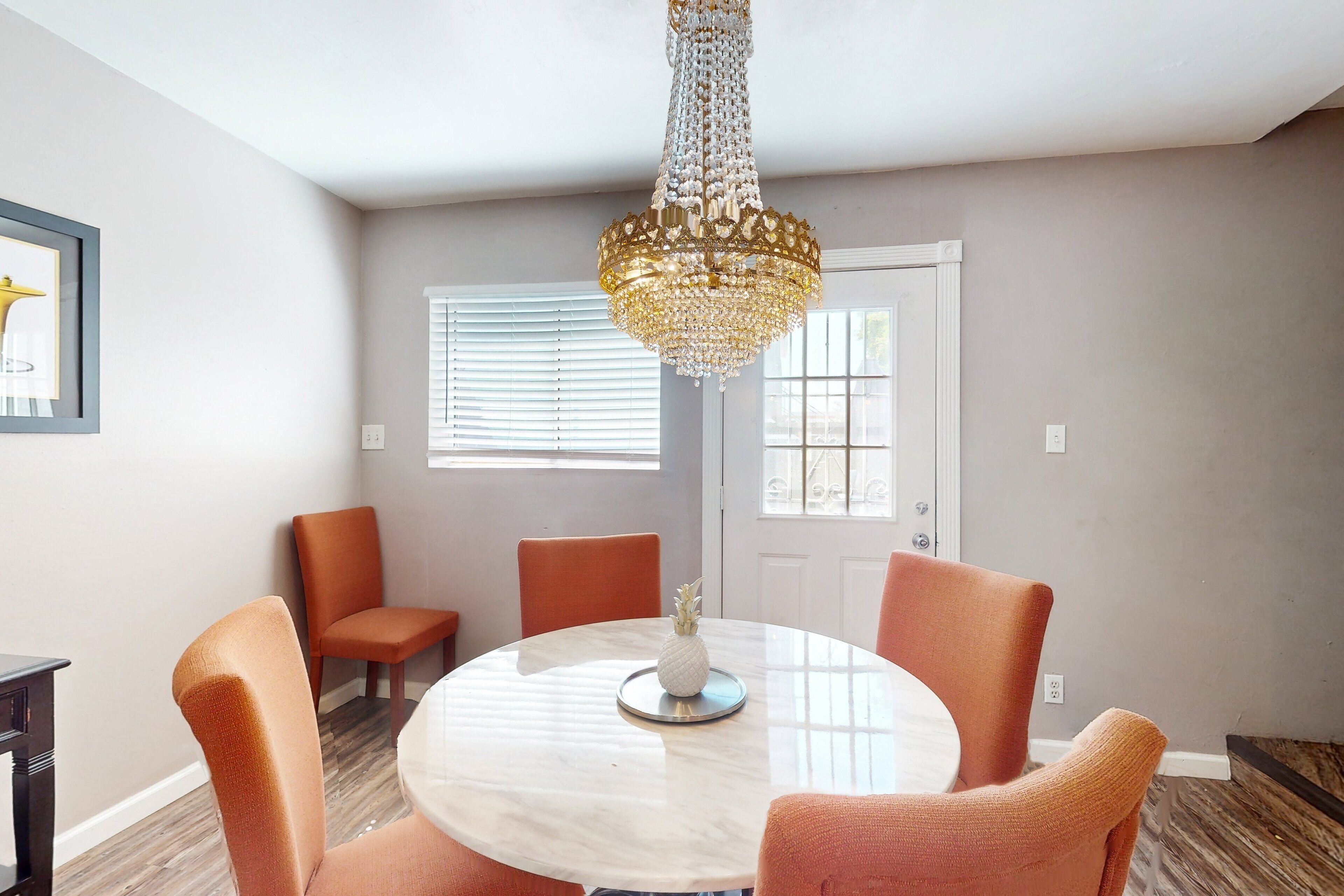 A dining area with a round table, orange chairs, a chandelier, and a framed picture on the wall.
