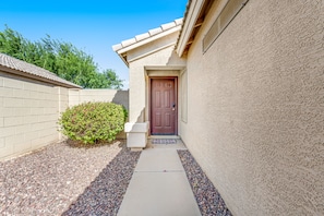 A narrow pathway leading to a door, flanked by a brick wall and a stucco wall, with a small bush and a concrete planter on the left. - Spacious home with private heated pool with waterfall, putting green & firepit (Goodyear)