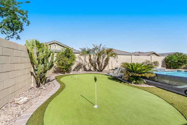 A backyard with a putting green, a pool, and a house.
