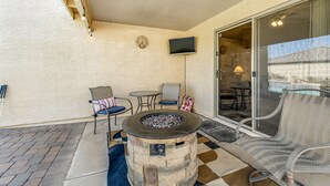 A patio area with a stone fire pit, outdoor seating, a television, and a view of a swimming pool.