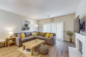 A living room with a sectional sofa, a coffee table, a side table, a lamp, a clock, a fireplace, and a window with blinds. - Desert Falls Condo on Golf Course w/Shared Fitness Center, Pools, Spas & Tennis (Palm Desert)