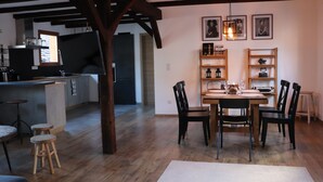 A dining area with a wooden table and chairs, a kitchen in the background, and wooden beams on the ceiling.