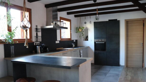 A modern kitchen with a central island, bar stools, a black refrigerator, and hanging plants.