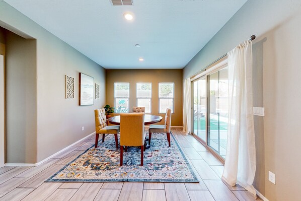 A dining area with a round table, chairs, a patterned rug, and a view through large windows.
