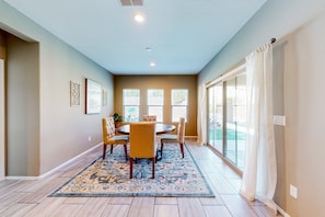 A dining area with a round table, chairs, a patterned rug, and a view through large windows.