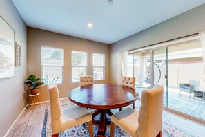 A dining area with a round wooden table surrounded by four cushioned chairs, large windows with blinds, a potted plant, and a view of the backyard through glass doors.