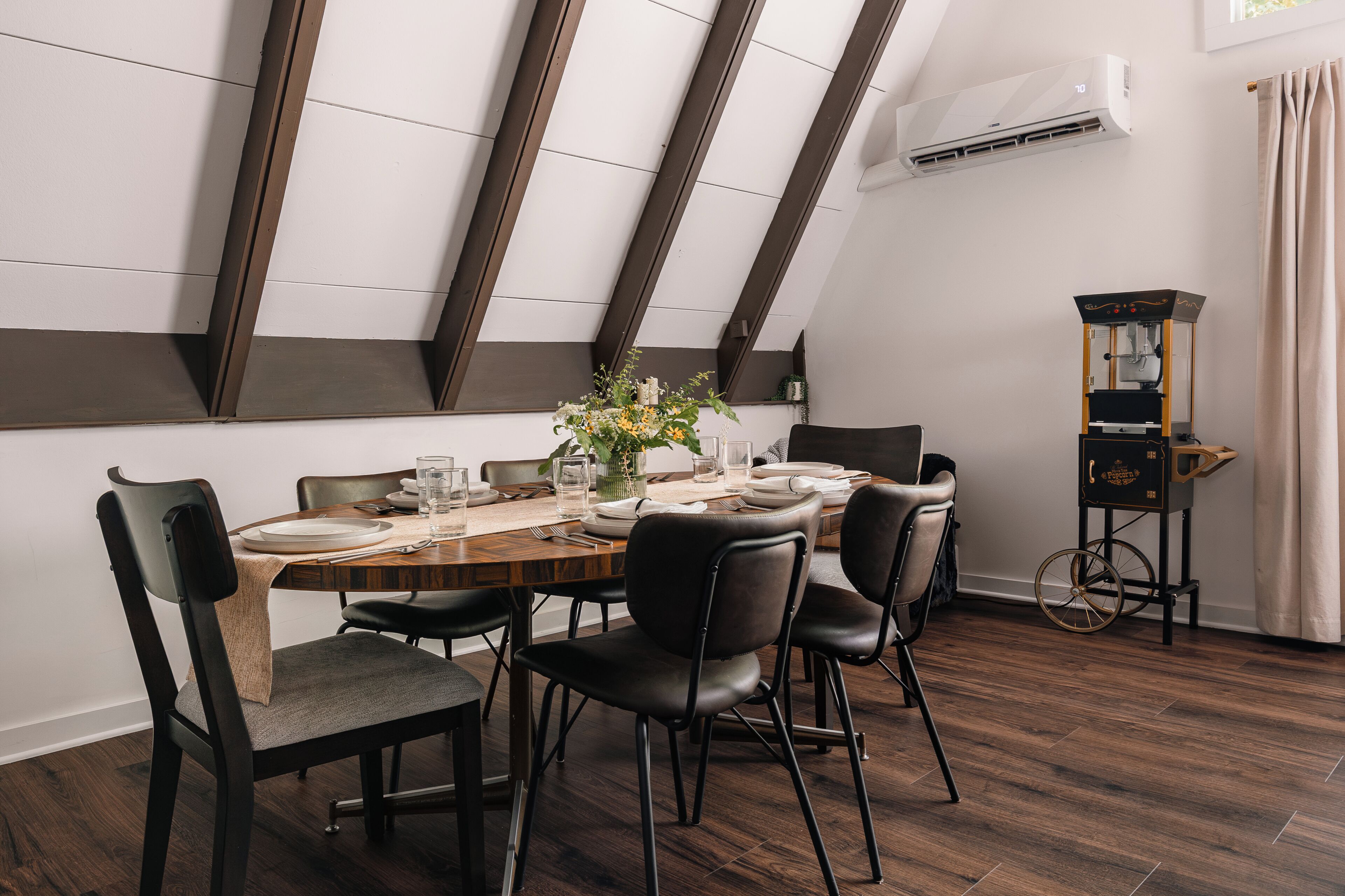 A dining area with a wooden table set for six, surrounded by black chairs, in a room with sloped ceiling and exposed beams.