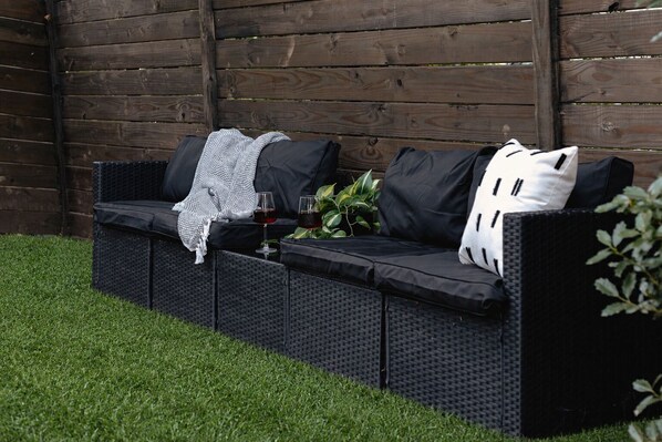 A black outdoor sofa with cushions and a blanket, accompanied by two glasses of red wine and a potted plant.