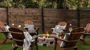 A fire pit surrounded by Adirondack chairs with blankets and candles, set against a wooden fence.