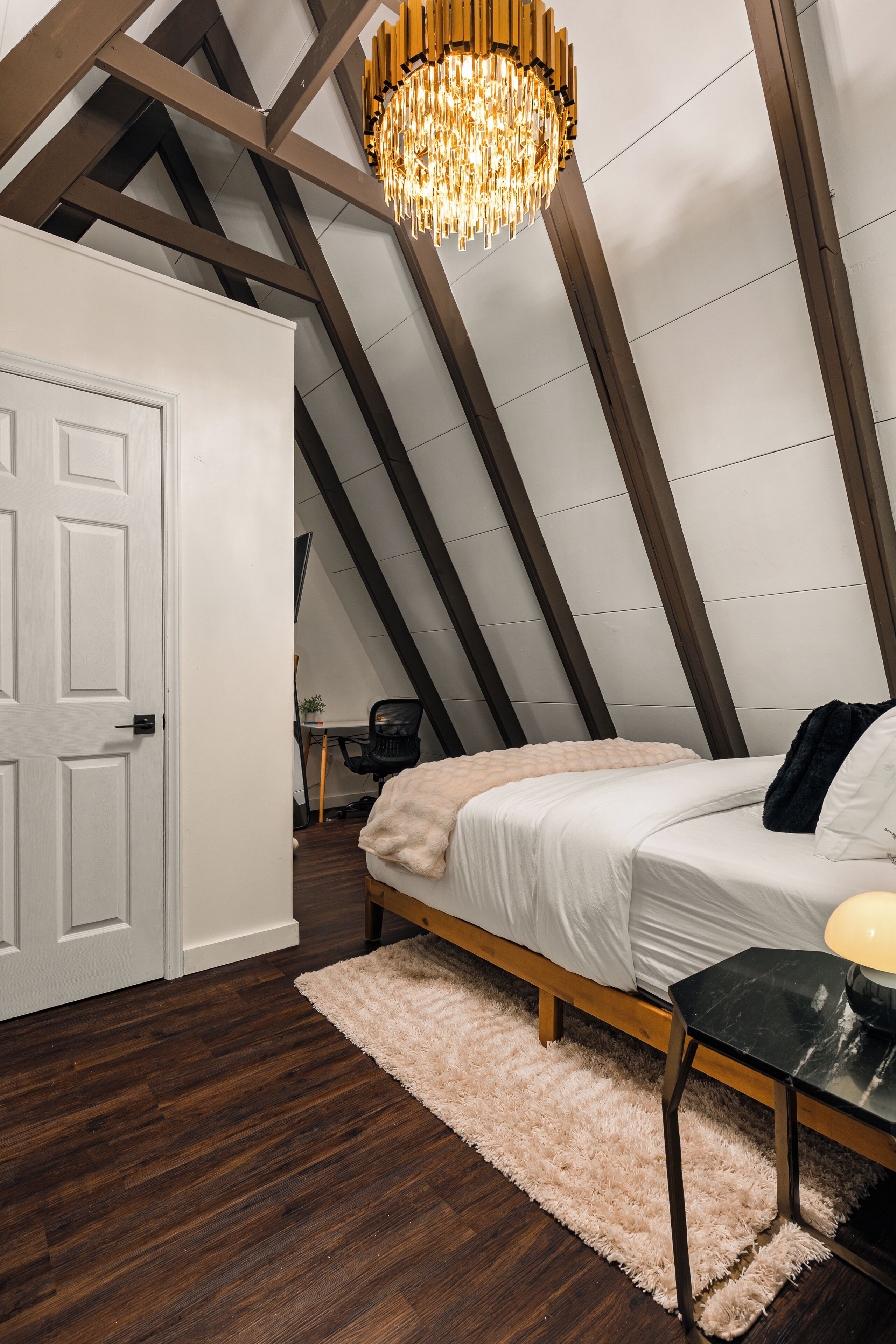 A bedroom with a sloped ceiling, a bed, a desk, and a chandelier.