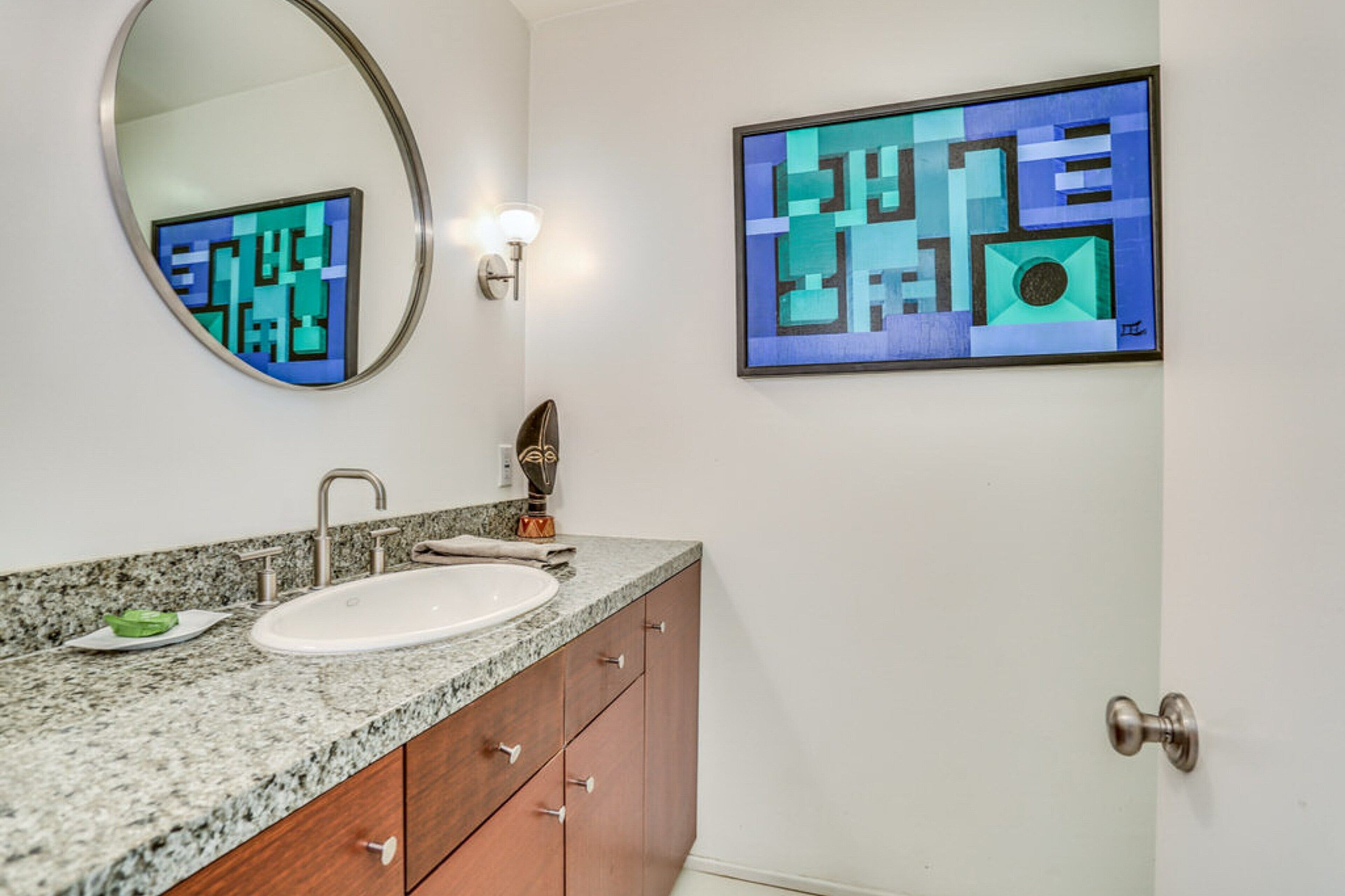 A bathroom with a granite countertop, a white sink, and a mirror. There is a framed abstract painting on the wall.