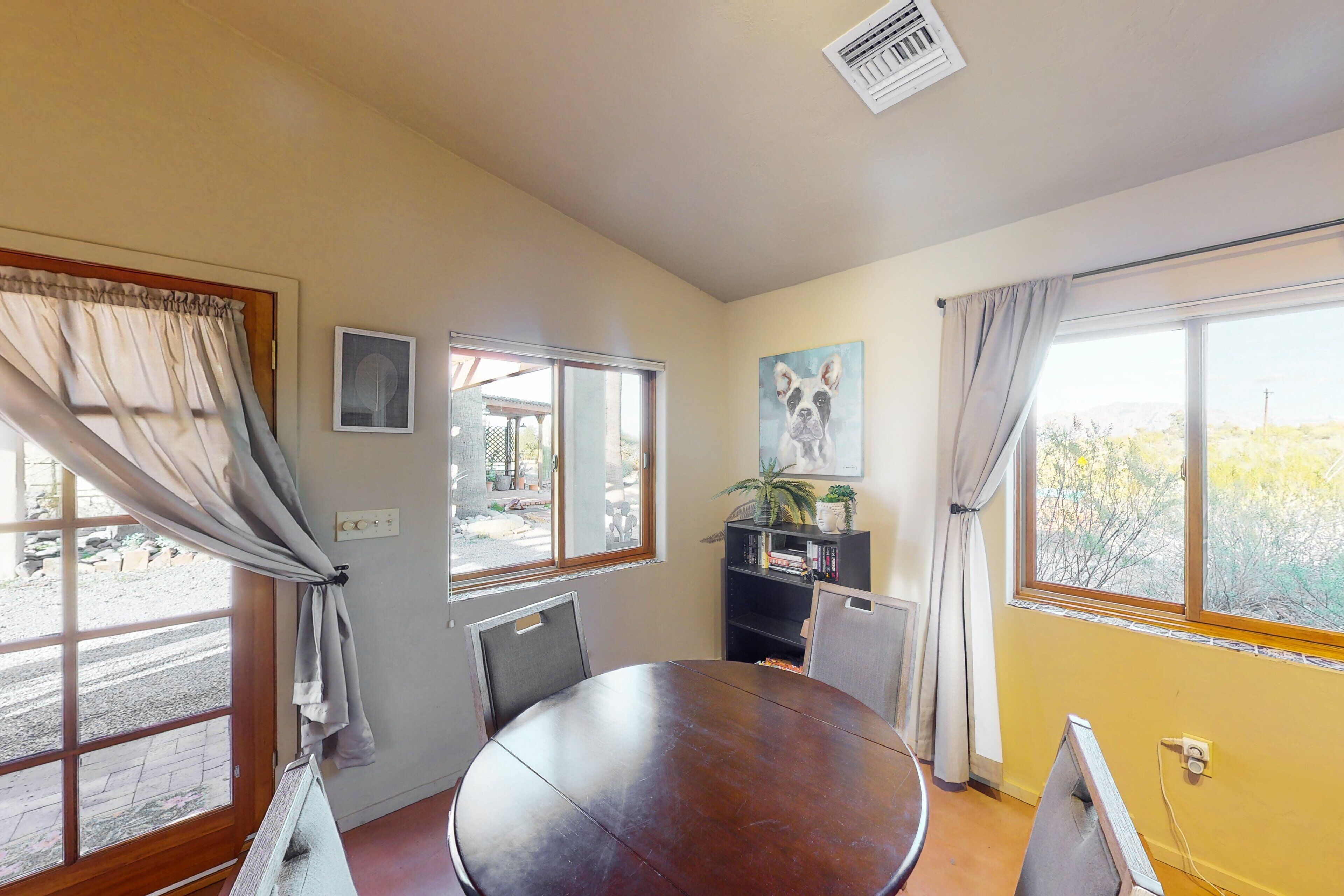A room with a round table, two windows with curtains, a radiator, and a bookshelf with a plant.
