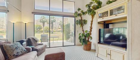 A spacious living room with a large TV, comfortable seating, a coffee table, and a large window offering a view of the outdoors.