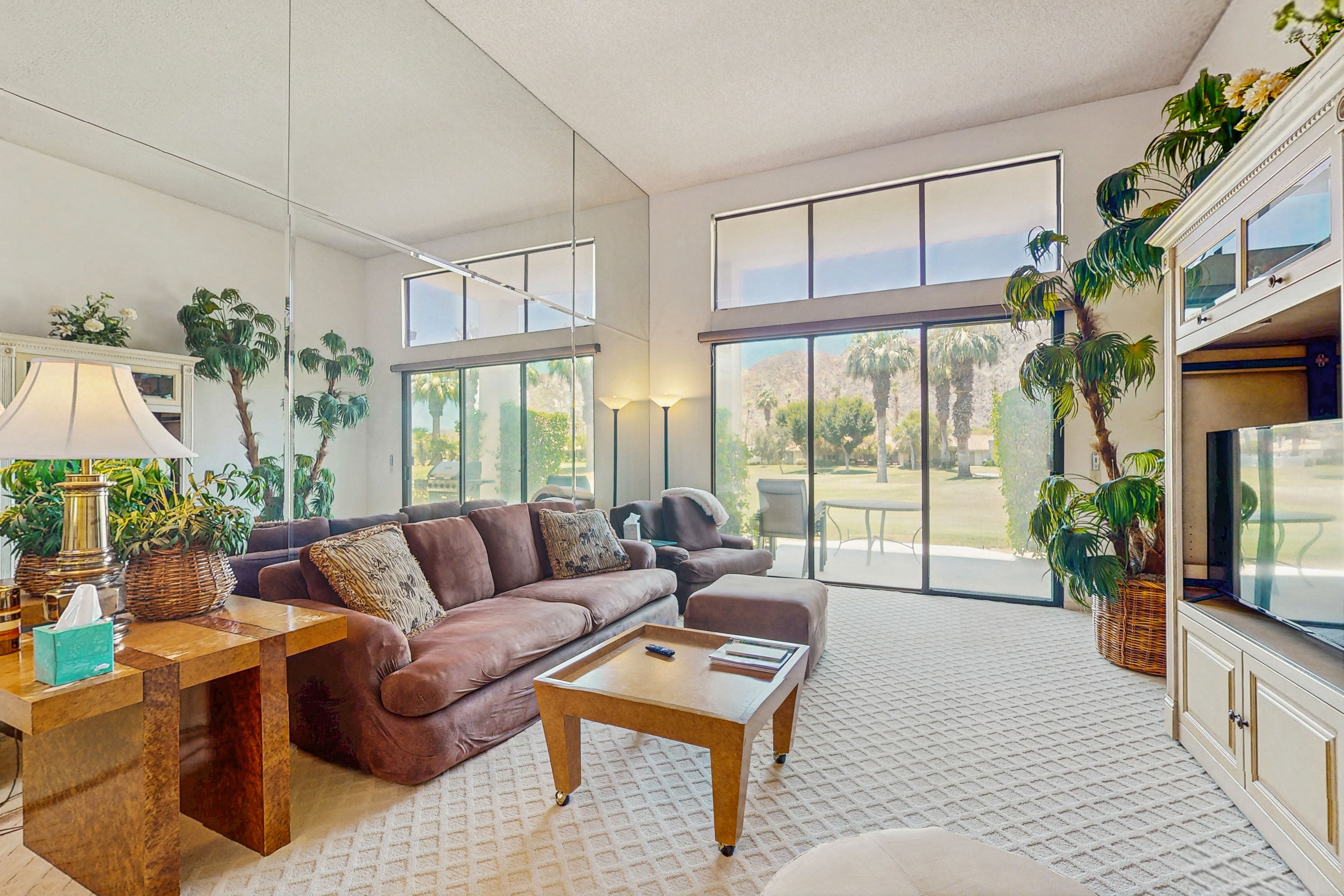 A spacious living room with a brown leather sofa, a wooden coffee table, and a large TV. There are several potted plants and a large window providing a view of the outdoors.