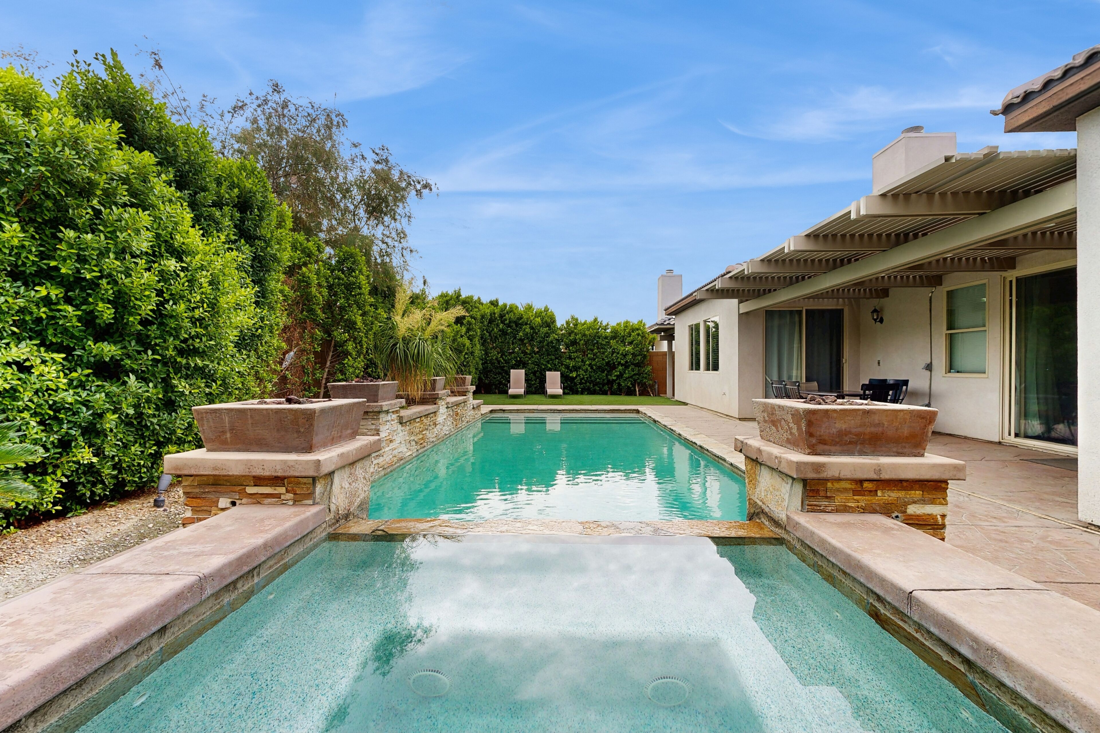 Ein rechteckiger Pool mit Steinstufen, umgeben von üppiger Vegetation und einem modernen Haus mit überdachter Terrasse.