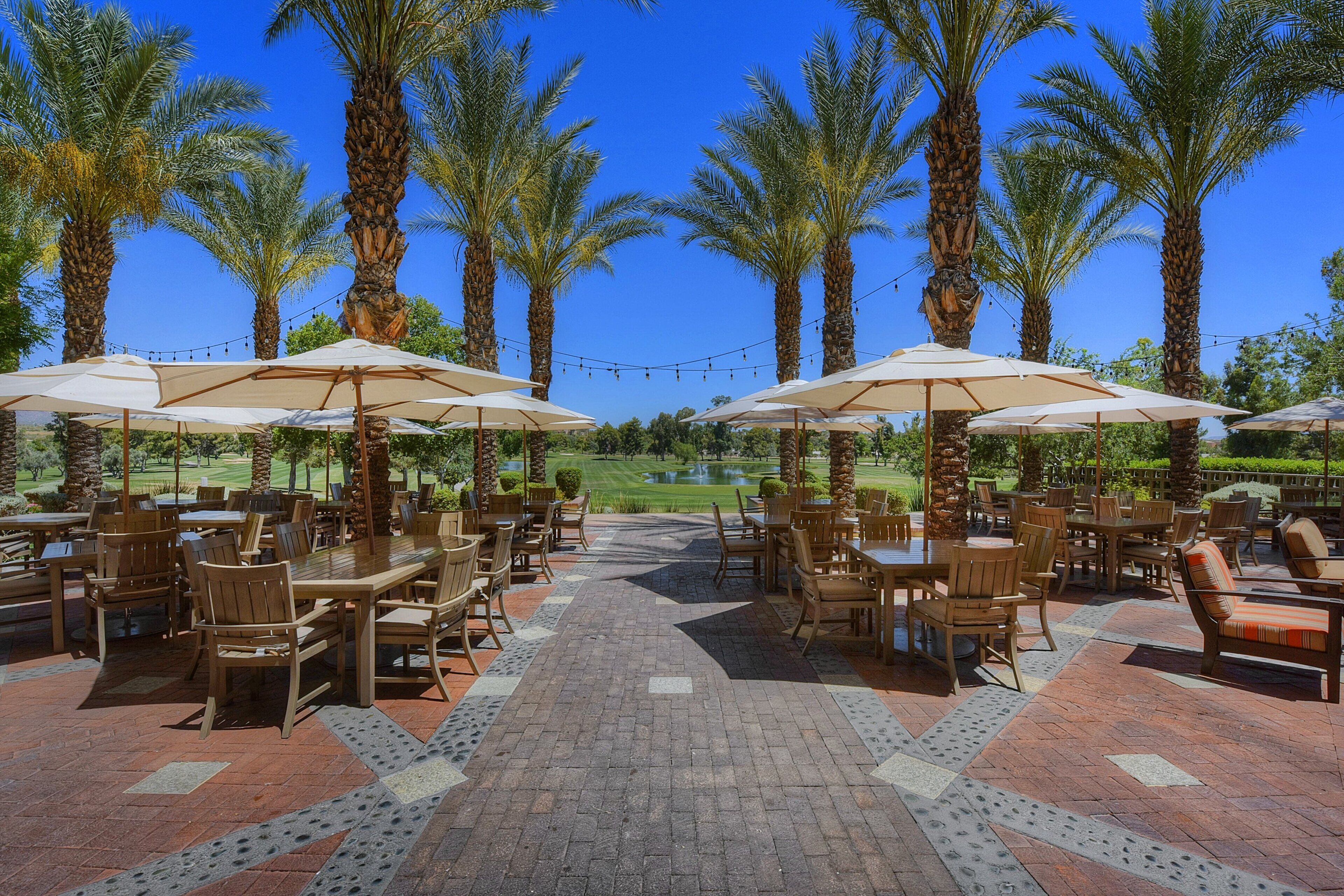 An outdoor patio area with tables and chairs, surrounded by palm trees, under clear blue skies.