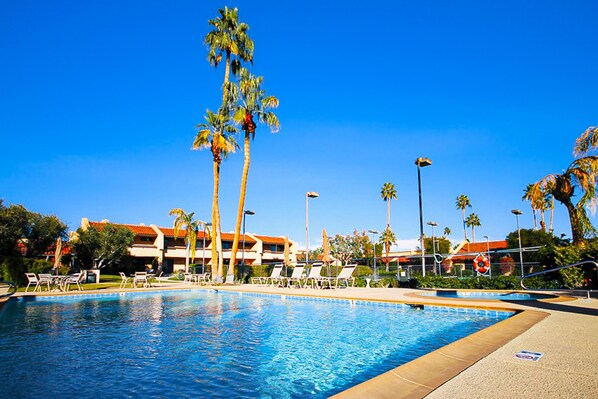 Una piscina con sillones de descanso y palmeras, rodeada de edificios con tejados rojos.