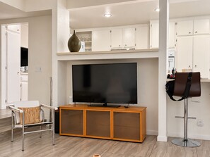 A modern living room with a flat-screen TV, a wooden cabinet, a leather bag on a stand, and a chair with a patterned cushion. - Dog friendly condo w/two patios & shared pool/tennis - walk downtown (Palm Springs)