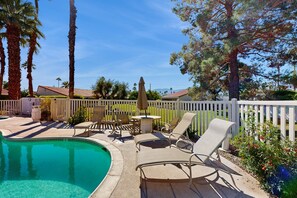 A pool area with lounge chairs and a table, surrounded by a white fence and palm trees. - Dog-Friendly Updated Condo Next to Golf Courses w/WiFi, AC, W/D, Shared Pool (Palm Desert)
