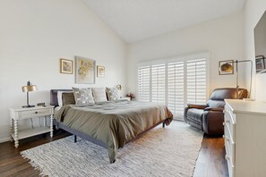Ein Schlafzimmer mit einem großen Bett, einem Nachttisch, einer Kommode, einem Sessel und einem Fenster mit Jalousien.