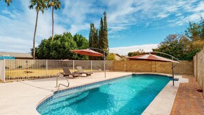 A swimming pool with a curved edge, surrounded by a fenced patio area with lounge chairs and umbrellas. - Welcoming home with private outdoor pool, pool table, patio, grill & central AC (Phoenix)