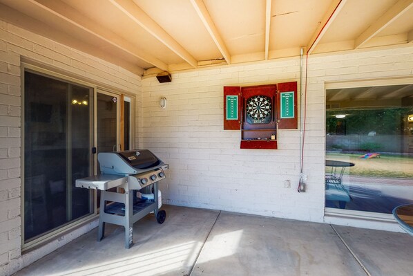Un patio couvert comprenant un barbecue, un jeu de fléchettes et une porte vitrée donnant sur un espace extérieur.