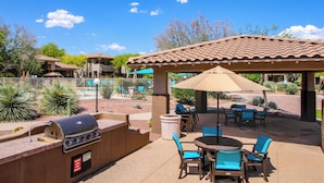 Un patio exterior con una parrilla, sillas y un área cubierta con una sombrilla.