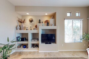 A living room with a flat-screen TV, shelves with decorative items, and a window with blinds. - Comfortable, updated condo with pool & hot tub (Oro Valley)