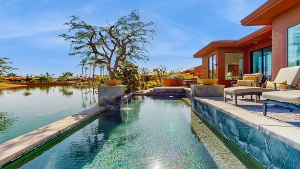 A swimming pool with a waterfall feature, surrounded by outdoor seating and a large tree. - Lakeside luxury home w/ private pool & spa, fireplace & outdoor kitchen-dog ok (La Quinta)