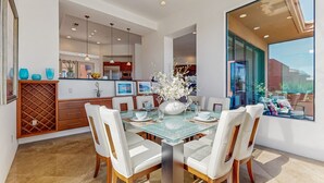 A modern dining area with a glass table, white chairs, and a kitchen in the background. - Lakeside luxury home w/ private pool & spa, fireplace & outdoor kitchen-dog ok (La Quinta)