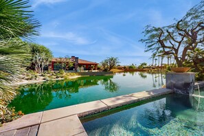 A swimming pool with a stone edge, surrounded by tropical plants and a building with a red roof. - Lakeside luxury home w/ private pool & spa, fireplace & outdoor kitchen-dog ok (La Quinta)