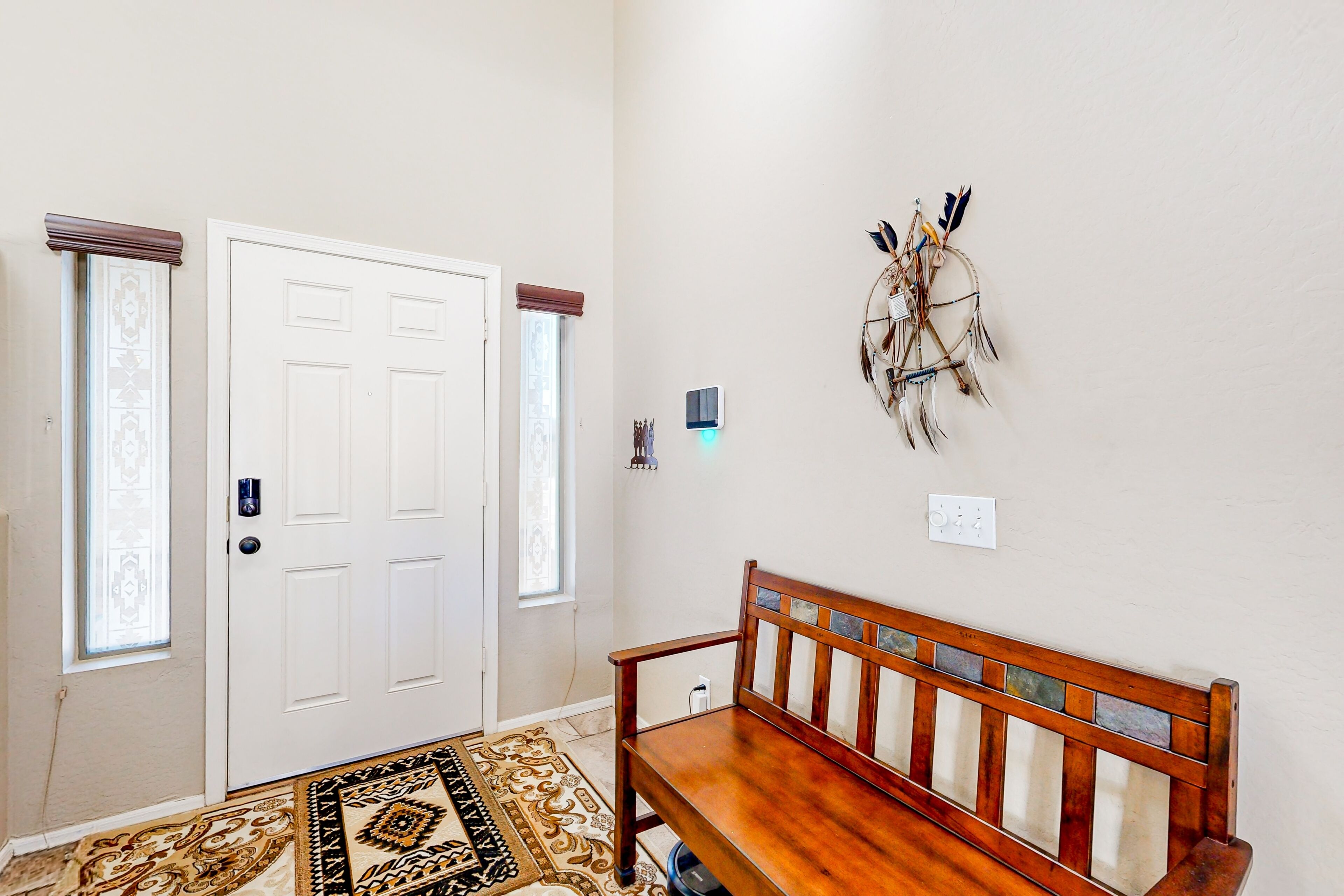 A hallway with a white door, a bench, a patterned rug, and a dreamcatcher on the wall.