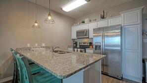 A kitchen with granite countertops, stainless steel appliances, and teal bar stools. - Ocean's 96 by Avantstay Heated Pool + Foosball Table + Media Room + Walk to North Padre Beach (Corpus Christi)