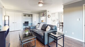 A modern living room with a dark leather sofa, glass coffee table, and a small side table. There is a kitchen area in the background with white cabinets and a dining table. - Golf-course-front condo near Old Town with two patios, WiFi, pool & hot tub (Scottsdale)