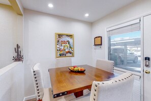 Un coin repas avec une table en bois, des chaises à revêtement blanc, un panier de fruits, un tableau encadré et une fenêtre avec des stores.