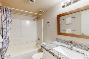Une salle de bain comprenant une baignoire, des toilettes, un lavabo avec un plan de travail en granit, une pomme de douche et un miroir avec un porte-serviettes.