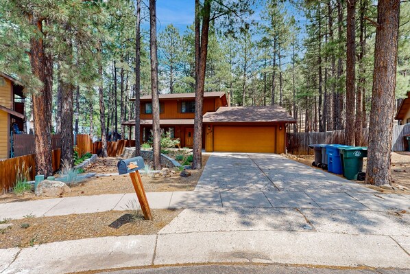 A two-story cabin with a stone driveway, surrounded by tall pine trees. - Lavish retreat with game room, tranquil porch & scenic yard-near skiing & dining (Flagstaff)