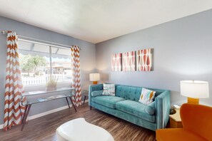 A room with a blue sofa, a white chair, a small table, and a window with curtains. - Snowbird-Friendly Condo with Patio Access, Central AC, Free WiFi - Dogs Welcome (Glendale)